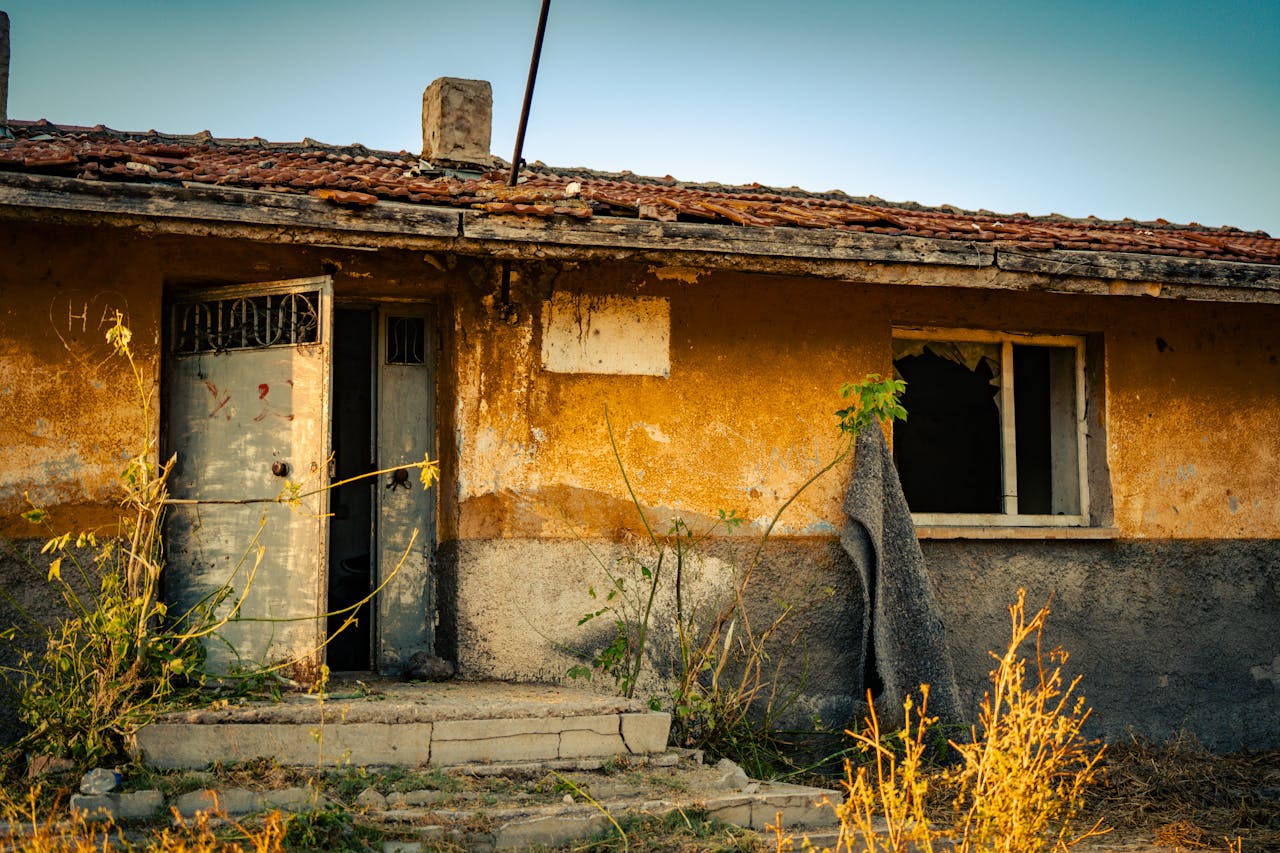 Weathered, abandoned house with broken windows and overgrown plants in a rural environment at dusk.